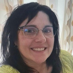 Frau mit Brille und langer, schwarzer Haar steht vor einem hellen Hintergrund. Die Person lächelt freundlich.